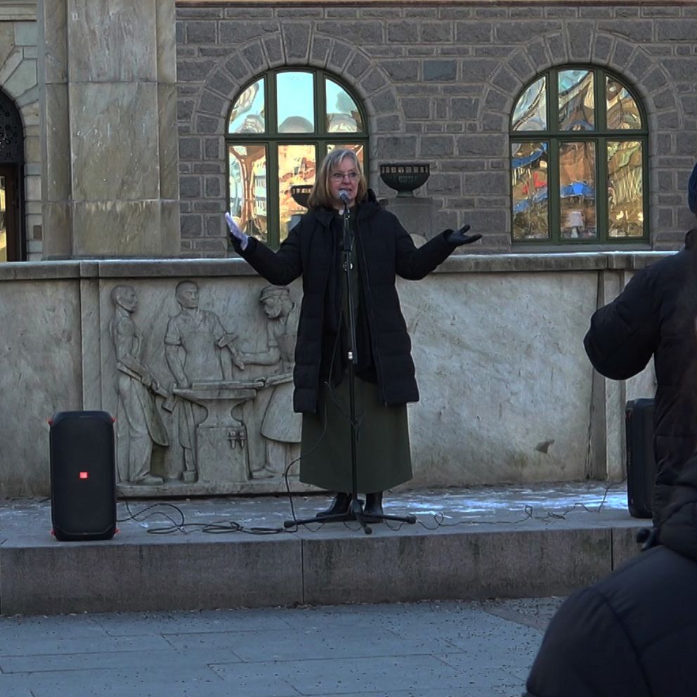 En person står på en trottoar och talar inför en liten publik utomhus. Personen är klädd i en mörk kappa och lång kjol och pratar i en mikrofon, med en byggnad i sten som bakgrund. Flera åhörare syns delvis i förgrunden.