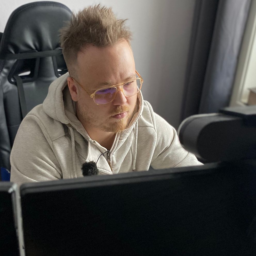 En person sitter framför flera datorskärmar i ett hemmakontor, iklädd en grå tröja. Rummet har en modern datorstol och ett fönster med gröna växter på fönsterbrädan.