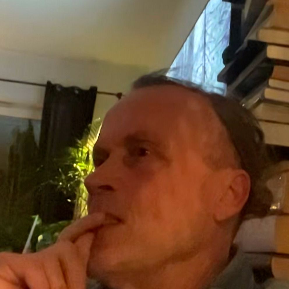 En man sitter i ett rum med en hög av böcker på hyllan i bakgrunden och en ljusgardin som släpper in utomhusljus.