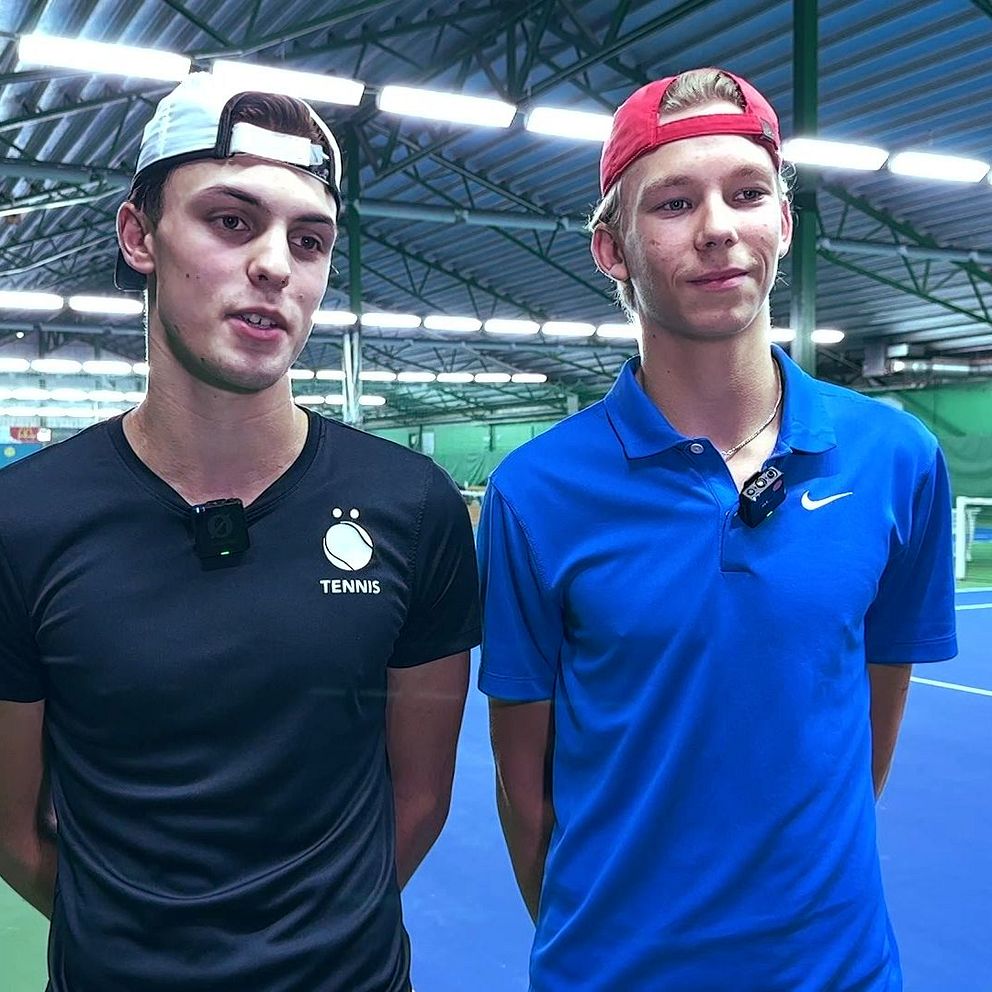 Två personer står bredvid varandra på en tennisplan inomhus. Den ena bär en svart t-shirt med texten ”TENNIS” och den andra en blå t-shirt med en Nike-logotyp. I bakgrunden syns en tennisspelare som slår en boll mot nätet.