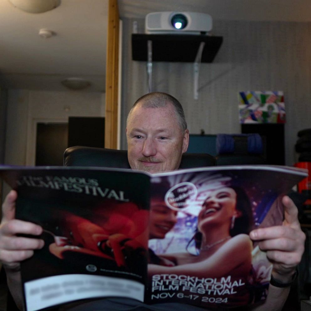 En person sitter i en mörk miljö och håller en broschyr för en filmfestival i händerna. På broschyren syns bilder relaterade till festivalen, inklusive en kvinna och text som anger datum för evenemanget, ”NOV 04-07 2024”. I bakgrunden syns en projektor och en lampa, vilket antyder en filmvisningssituation.