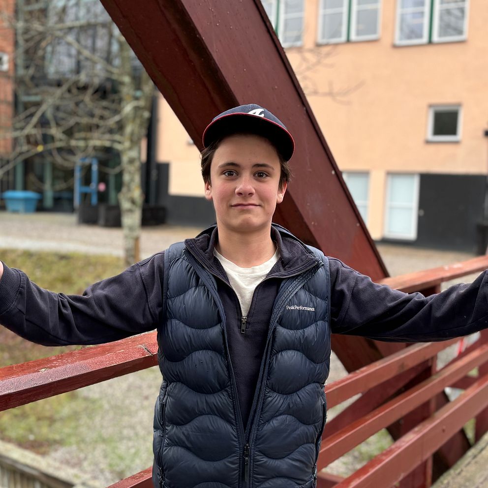 Person med keps står på bro med utsträckta armar, byggnader syns i bakgrunden.