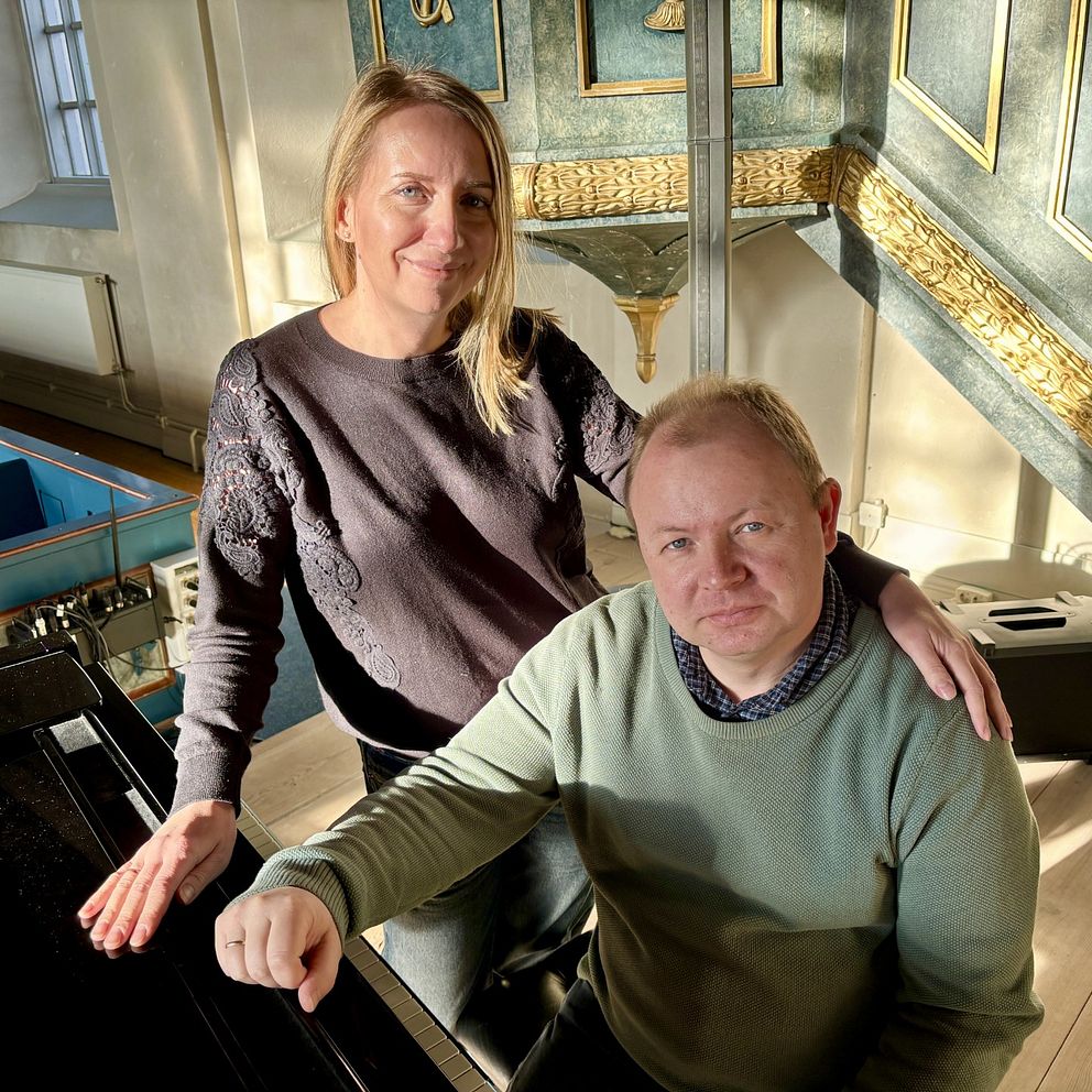 Två personer står intill varandra bredvid ett piano i ett rikt dekorerat rum med klassisk inredning och solljus som strömmar in genom fönstret.