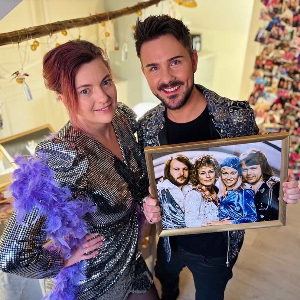 Två personer klädda i glittriga kläder håller i en inramad fotografi med Abba. De står i ett utrymme dekorerat med en vajer fylld med bilder och färgglada klädnypor i bakgrunden.