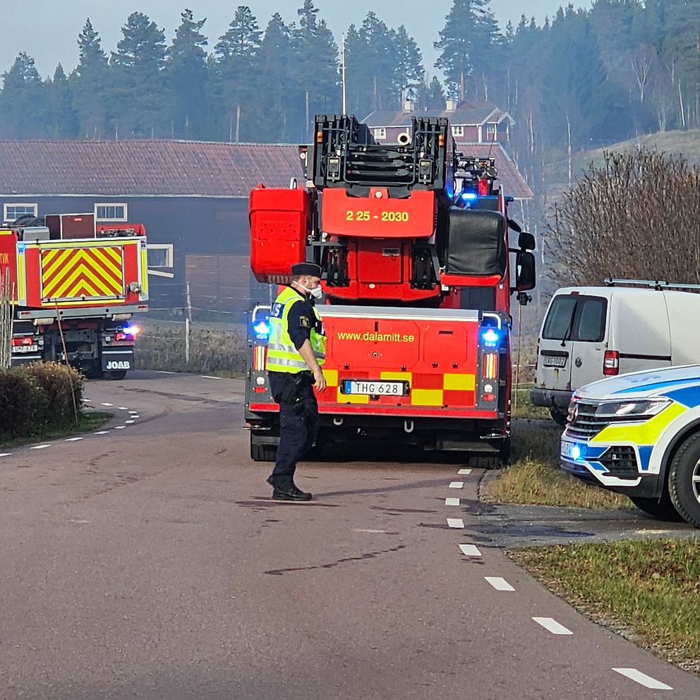 Brandbilar och en polisbil på en väg i Rättvik. En polisman med skydd för ansiktet står på vägen och rök syns i luften.