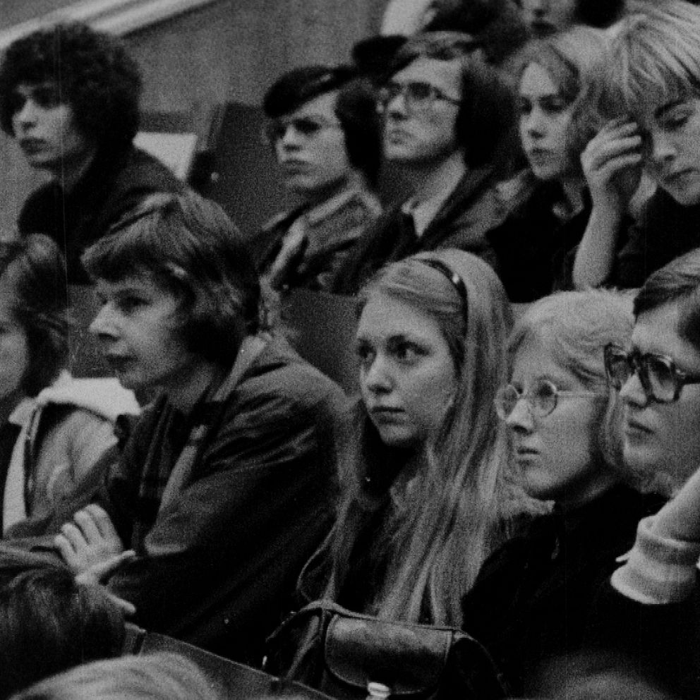 En grupp människor sitter i en auditoriumliknande miljö, fokuserade framåt mot en presentation eller ett tal. Det finns både män och kvinnor i olika åldrar, klädda i 1970-talsmode, vilket ger en känsla av en historisk kontext.