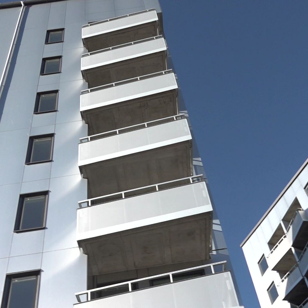 En bild av moderna flerfamiljshus med flera våningar i Umeå nära campus. Den vänstra byggnaden har balkonger i betong och en reflekterande fasad, medan den högra byggnaden har en mer traditionell design med stora fönster. Himlen är klar och blå, vilket skapar en kontrast till byggnadernas silverfärgade yta.