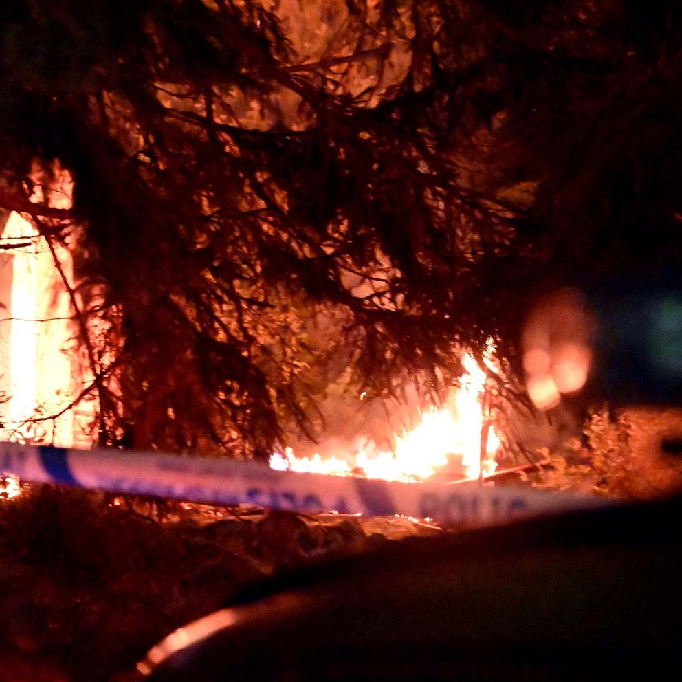Lågorna från en brand syns genom träden, medan en polisavspärrning markerar området. Bilden förmedlar en känsla av en pågående nödsituation under natten.