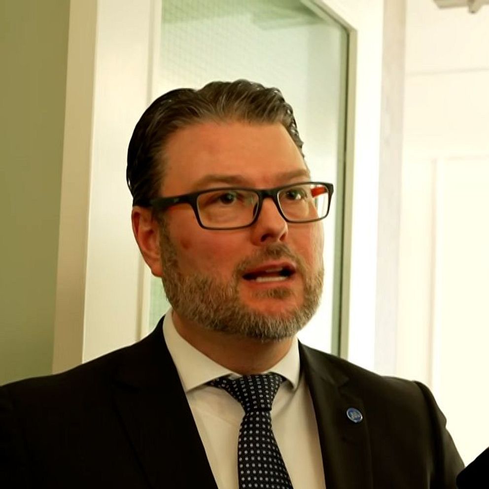 Edward Riedl (M) i kostym intervjuas av flera journalister som håller fram mikrofoner.
