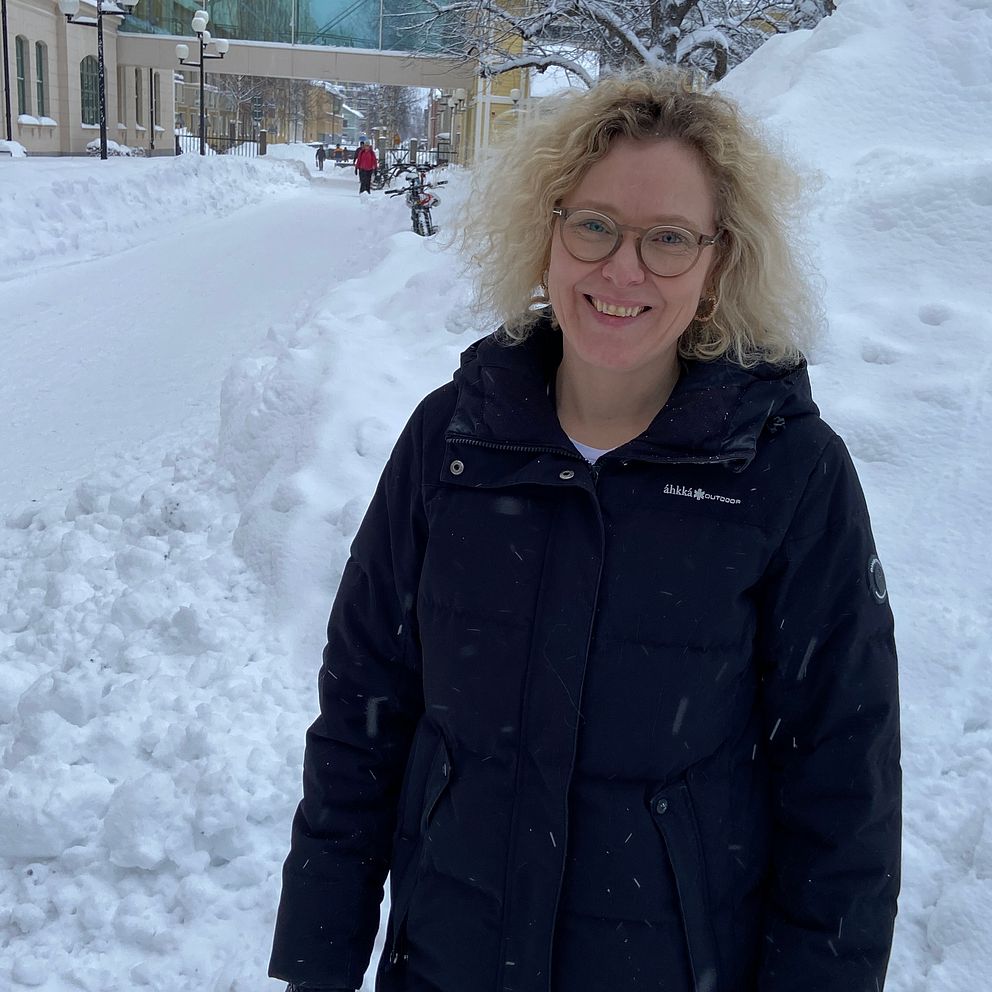 Personen står i en snöig stadsmiljö med höga snöhögar runt omkring. Gatan är täckt av snö och det är snöfall, vilket skapar en vintrig atmosfär.