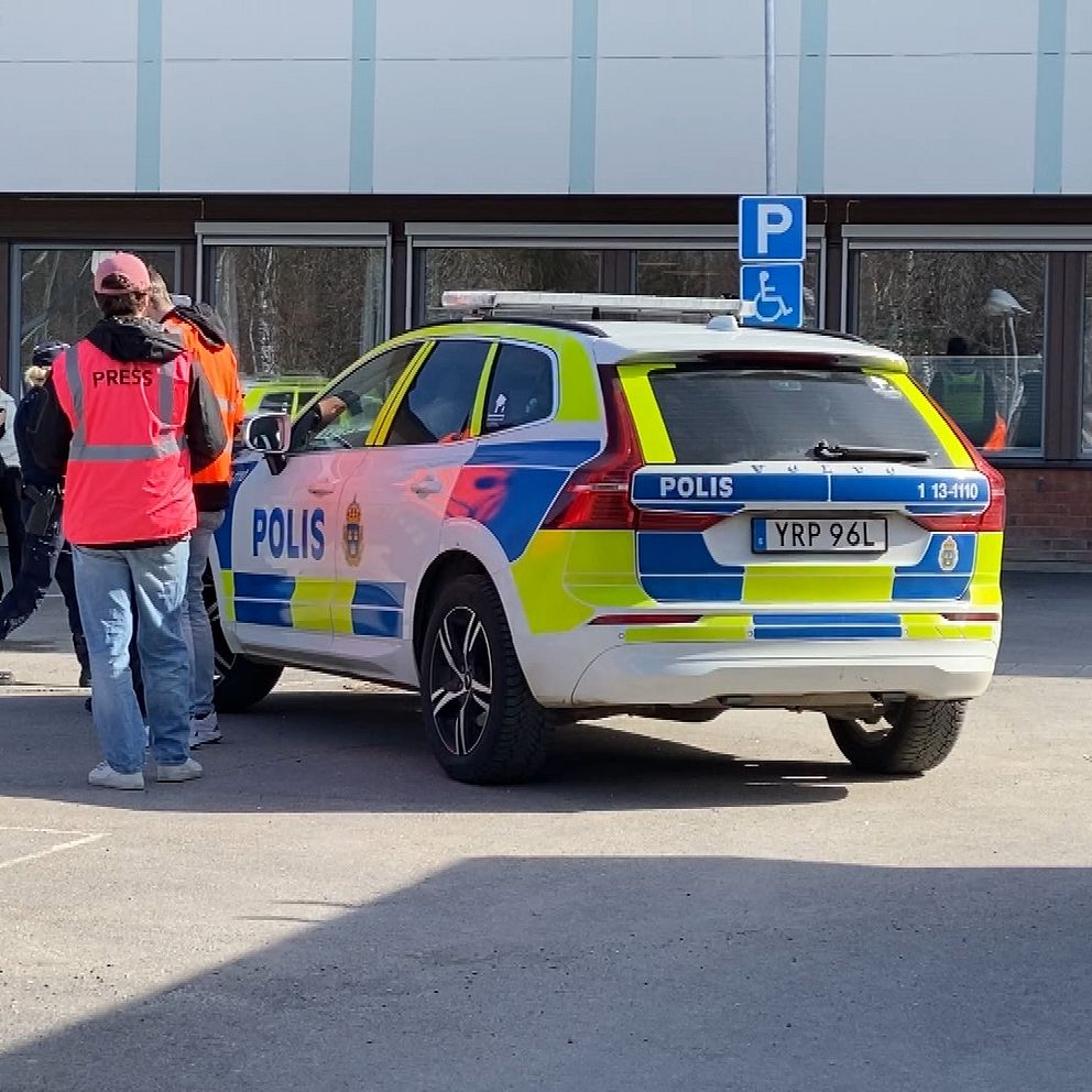Polisbil parkerad utanför en byggnad. Person i röd väst pratar med flera människor.