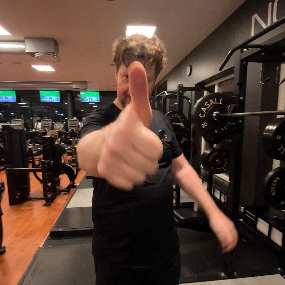 En person i träningskläder ger tummen upp på ett gym, med träningsmaskiner och vikter i bakgrunden.