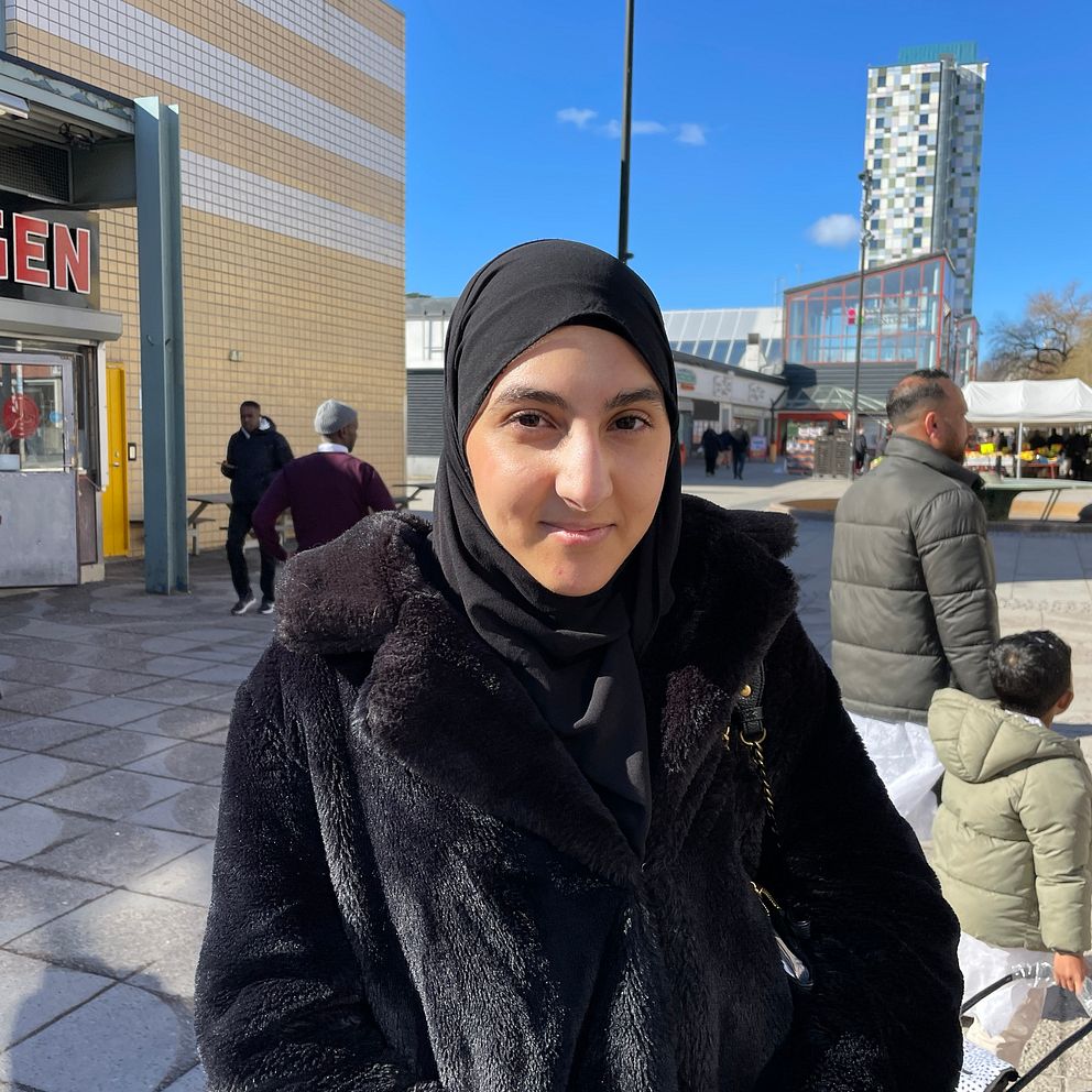 En ung kvinna står på Tensta torg i svart jacka och ler. Det går folk i bakgrunden.