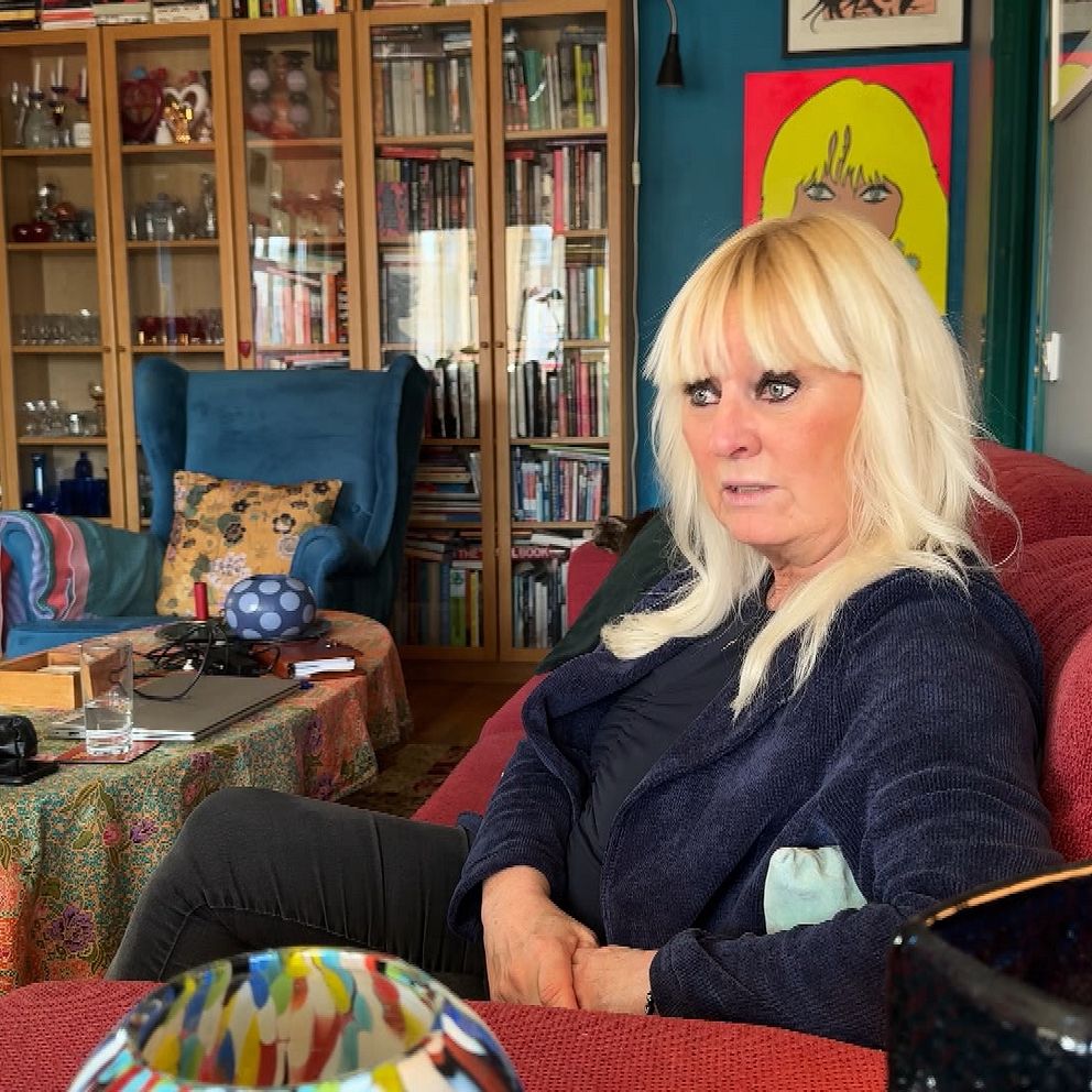 En kvinna sitter i ett rum fyllt med böcker och färgglada möbler, med en tavla synlig på väggen i bakgrunden. Miljön verkar personlig och hemtrevlig med olika objekt och en dekorativ duk på bordet.