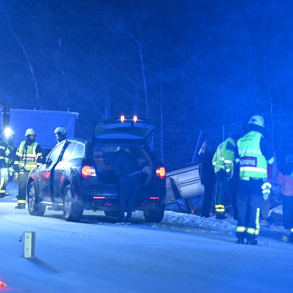 Räddningspersonal arbetar vid en bilolycka på en mörk väg, där en bil står förstörd vid sidan av vägen. Blåljus från en räddningstjänstbil bländar i bakgrunden, och flera brandmän och ambulanspersonal är synliga i sina uniformer, som undersöker situationen och hjälper skadade personer.