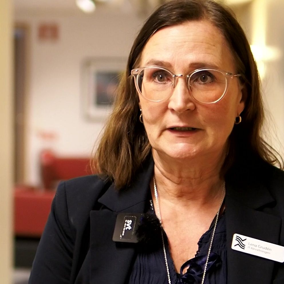 En person i en mörk kavaj med en namnbricka står i en korridor med beige väggar och röd soffa i bakgrunden. Namnbrickan och omgivningen tyder på en professionell miljö.