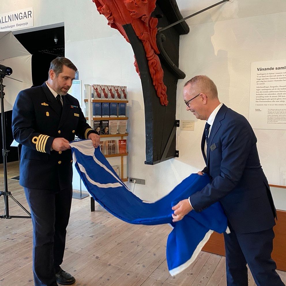 Två män i en formell miljö håller i en blå och vit flagga. Ena mannen är klädd i en uniform och den andra i kostym. De står bredvid en utställningsplats med en skylt i bakgrunden som säger ”Utställningar”.