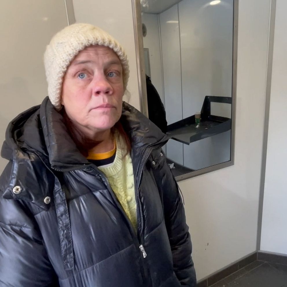 Petra Källberg med en mörkgrå täckjacka och vit stickad mössa är hemlös. Hon visar upp en offentlig toalett där hon brukar sova, väggarna är vita och det är en spegel i bakgrunden.