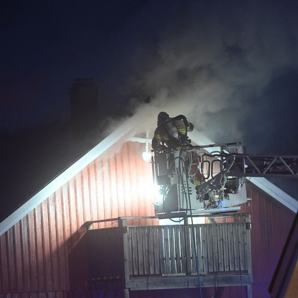 Brandmän bekämpar en brand på taket av ett hus, rök stiger upp i mörkret.