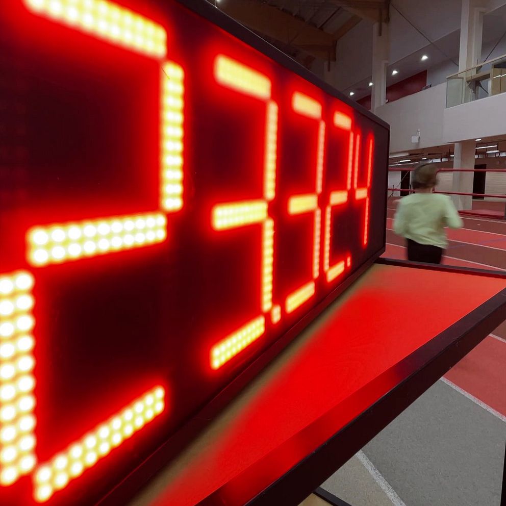 En stor, röd digital tidsdisplay som visar siffrorna ”23.324” i en inomhusidrottsanläggning. I bakgrunden syns en person som springer på en löparbana.