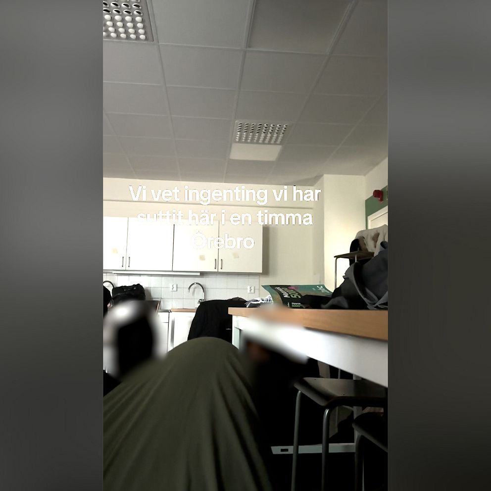 En person sitter i ett klassrum med vita skåp och bänkar. Texten över bilden lyder: ”Vi vet ingenting vi har suttit här i en timma Örebro”.
