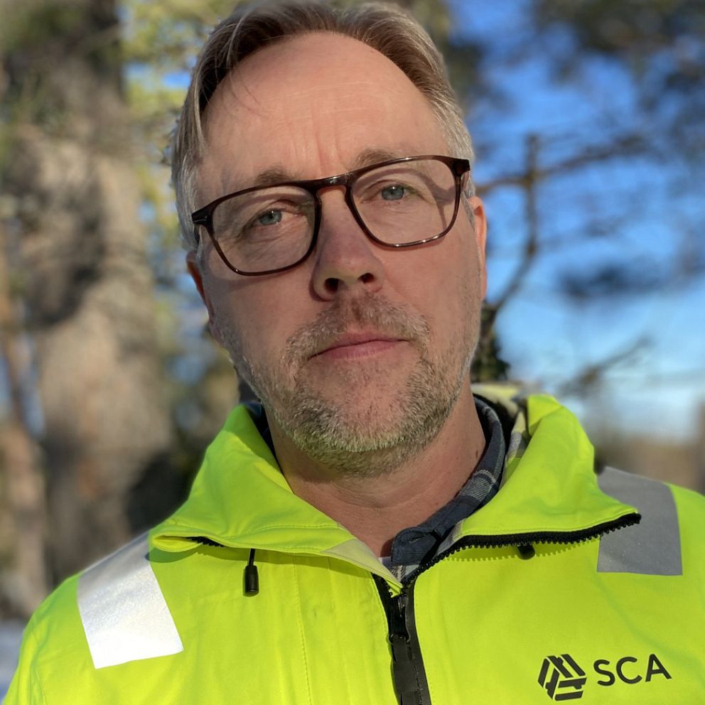 Person i gul SCA-jacka framför skogsmiljö under en solig dag.