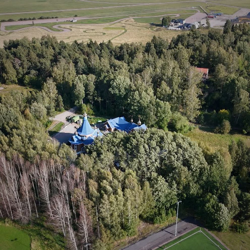 Flygfoto över ett hus med blått tak omgivet av tät skog och öppna fält.