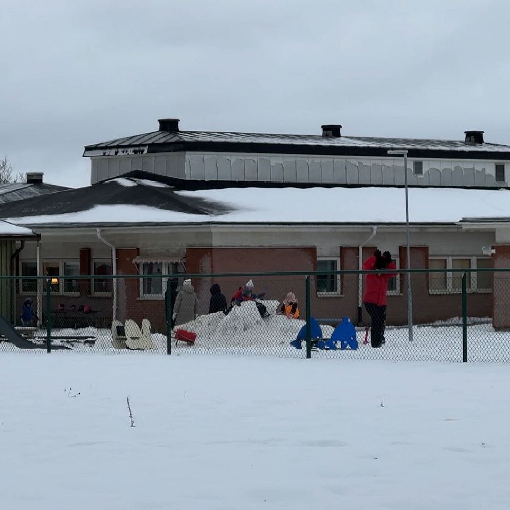 Barn leker utomhus i snön vid en förskola. I förgrunden syns ett lekställe med klätterställning och en snöhög där barn bygger. Byggnaden i bakgrunden är lågt belägen med takfönster och mörka fönster. Grå himmel och snö täcker marken.