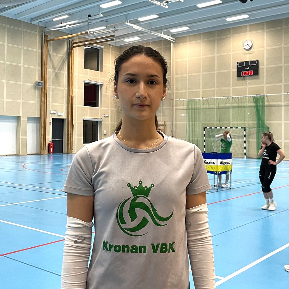 En ung kvinnlig volleybollspelare från Kronan VBK står i borstahusens idrottshall. Tränare och andra volleybollspelare syns i bakgrunden.