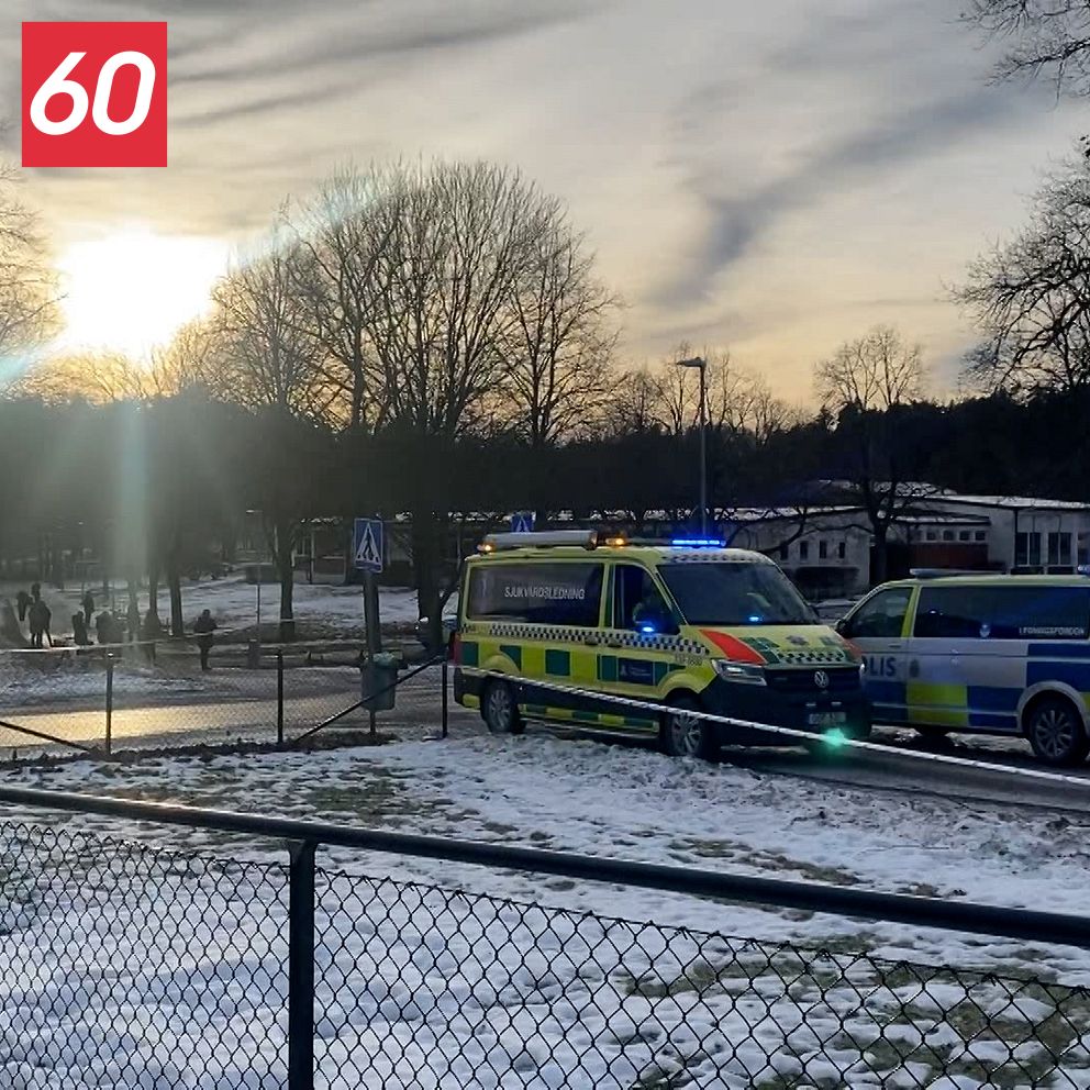 Två polisfordon står parkerade på en snötäckt gräsmatta, med solnedgången i bakgrunden. En del av en park är synlig med träd och människor i bakgrunden.