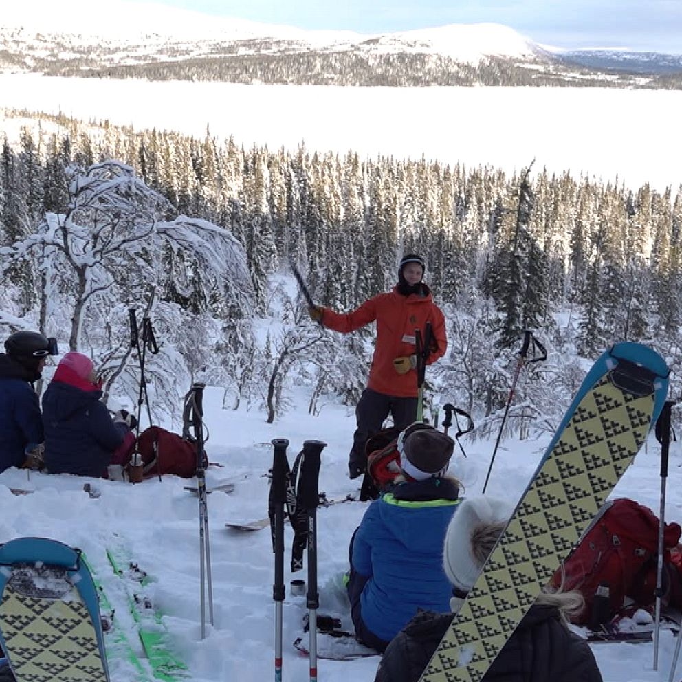 En grupp skidåkare sitter i snön och lyssnar på en guide som pratar framför dem, med en snöklädd skog och berg i bakgrunden. Vintrig landskapssceneri med skidutrustning synlig omkring dem.