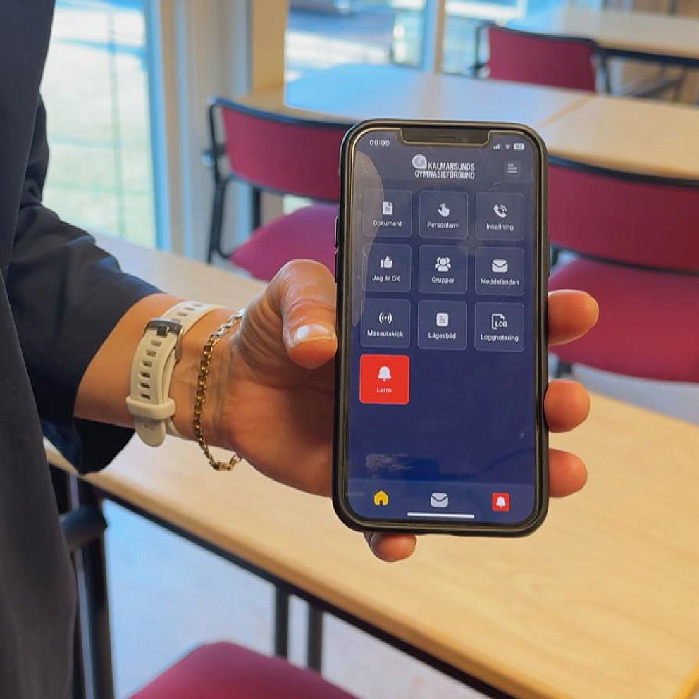 En person håller en smartphone med en säkerhetsapp öppen, där ett rött larm visas. I bakgrunden syns ett tomt klassrum med träbord och röda stolar.