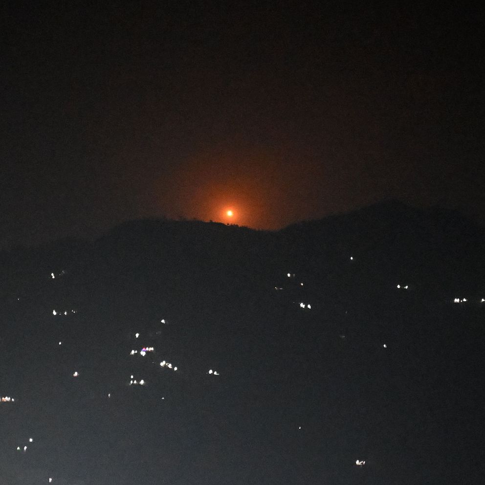 Enkel bild. Svart himmel med en orange ljusglimt bakom mörka bergstoppar på natten.