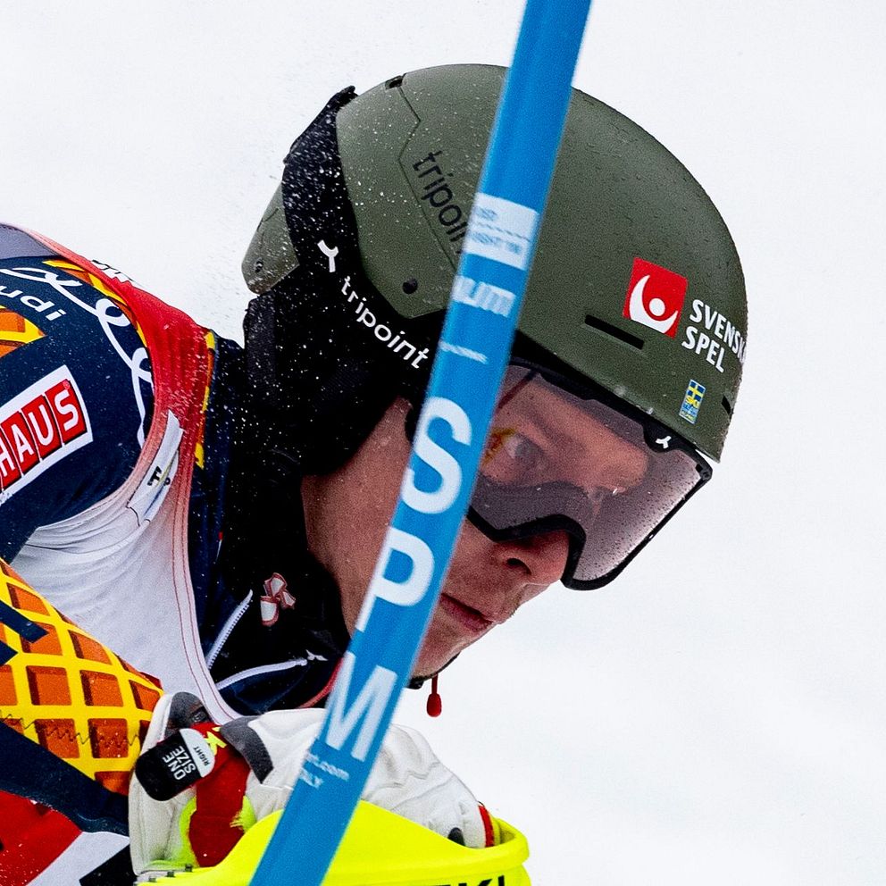 En alpin utförsåkare i tävlingsdräkt från Sverige navigerar genom en slalombana med markerade blå portstolpar, iklädd hjälm och med företagslogotyper tydliga på utrustningen.