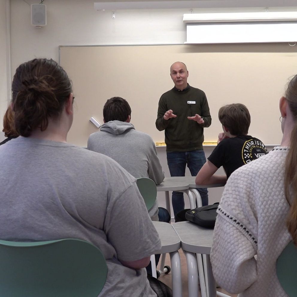 En lärare står framför en klassrumsgrupp av studenter som sitter vid sina bänkar, lyssnar och tittar framåt. Bakgrunden visar en vit tavla på väggen.