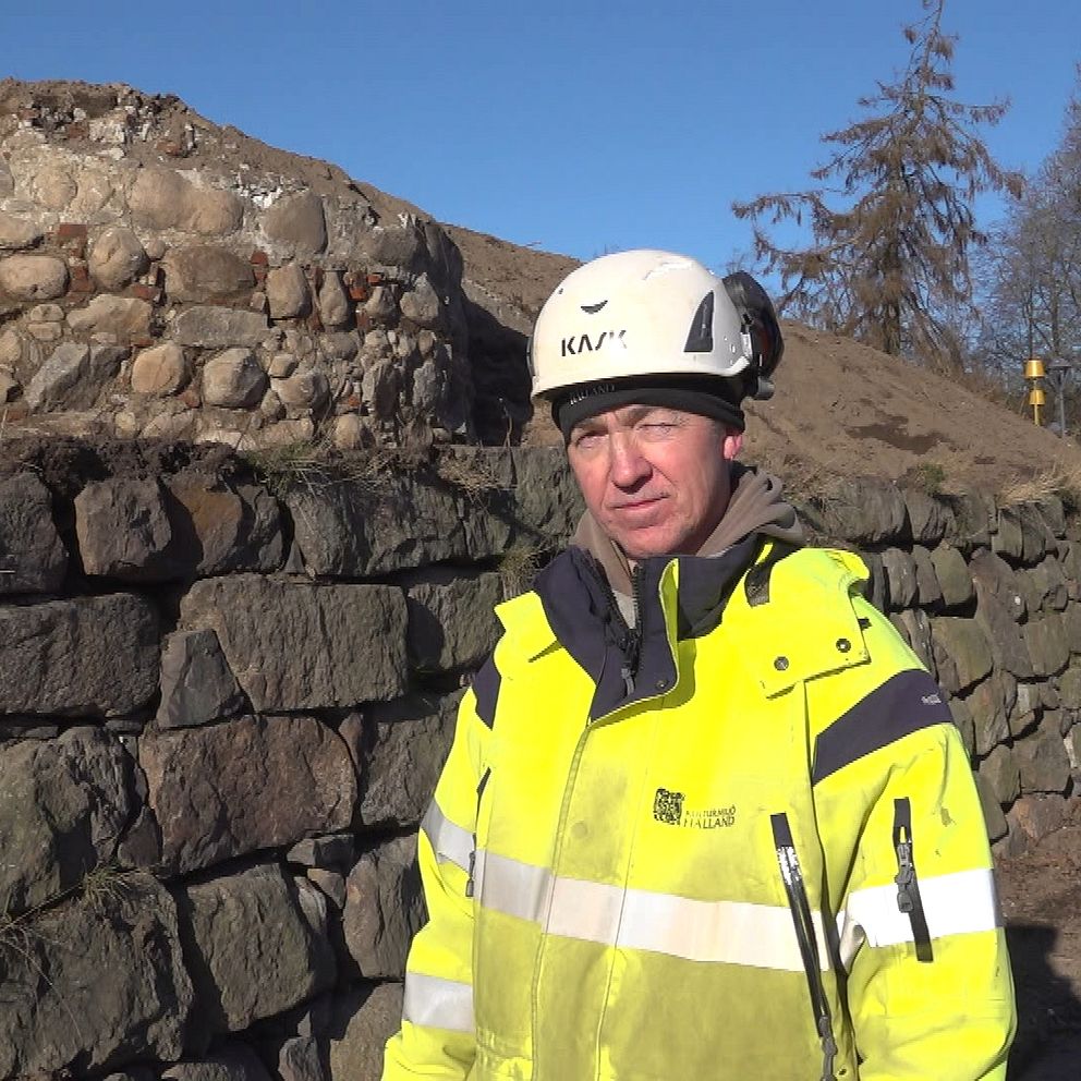 En person i varselkläder och hjälm står framför en gammal stenmur på en solig byggarbetsplats med träd i bakgrunden.