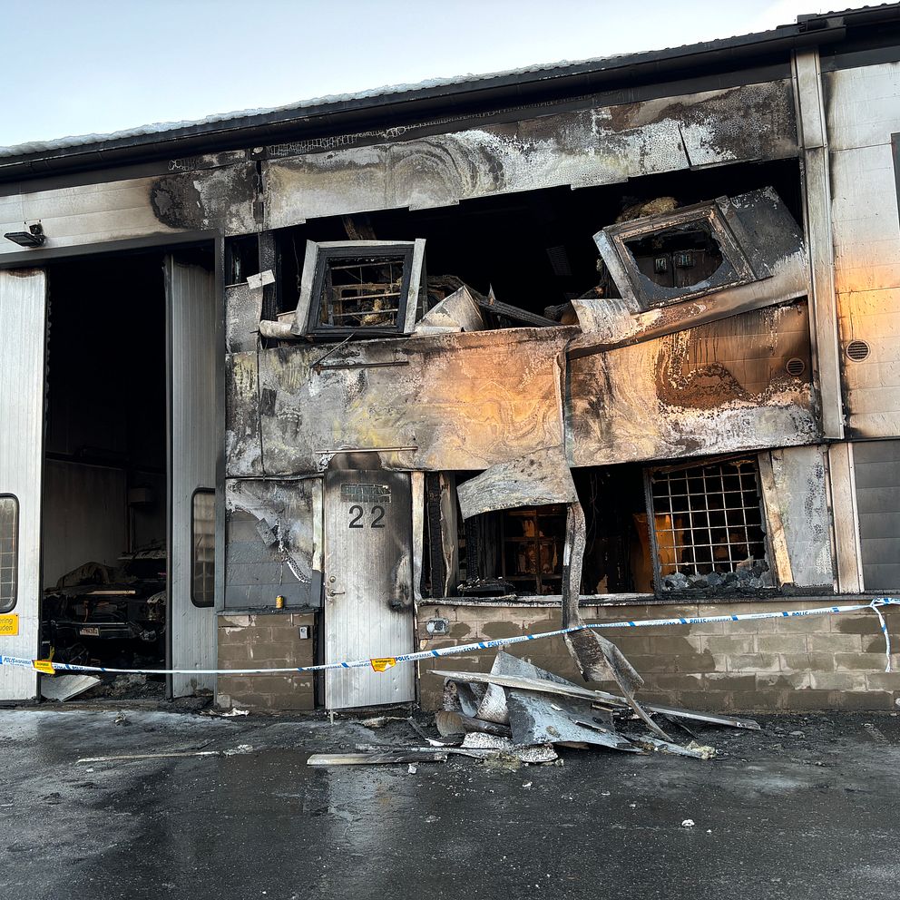 Skadat garage med omfattande brandskador, med förstörda dörrar och brända väggar. Numret 22 syns tydligt på dörren. Polisens avspärrning syns runt området.