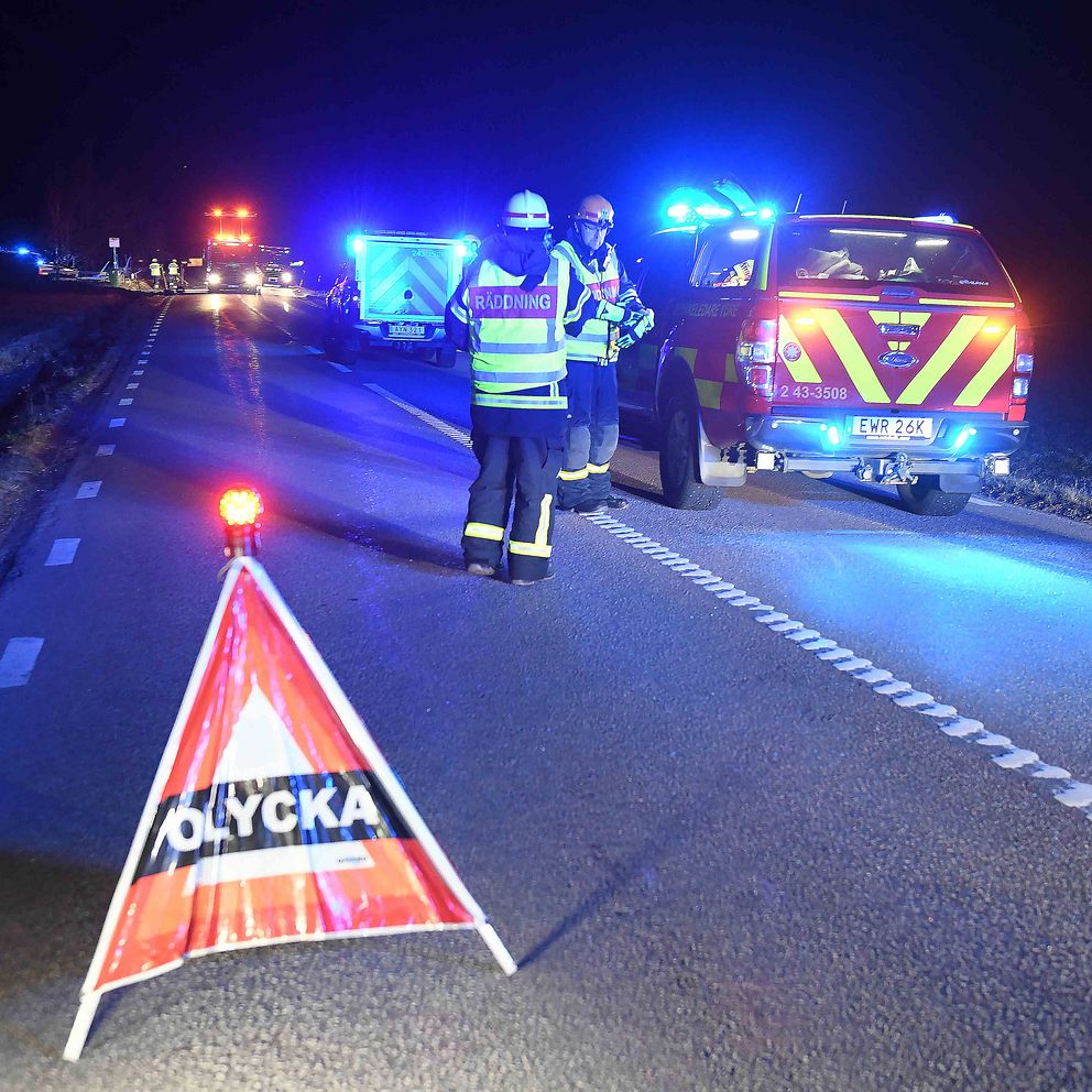 Utryckningsfordon och personal vid olycksplats på en väg under natten. Olycka-skylt i förgrunden.