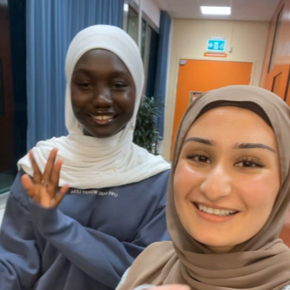 Två personer står i en inomhusmiljö, båda klädda i hijab. Den första personen, till vänster, har på sig en ljusblå tröja och gestikulerar med handen. Den andra personen, till höger, bär en beige hijab och ler. Bakgrunden innehåller inredning med blå väggar och fönster som släpper in ljus.
