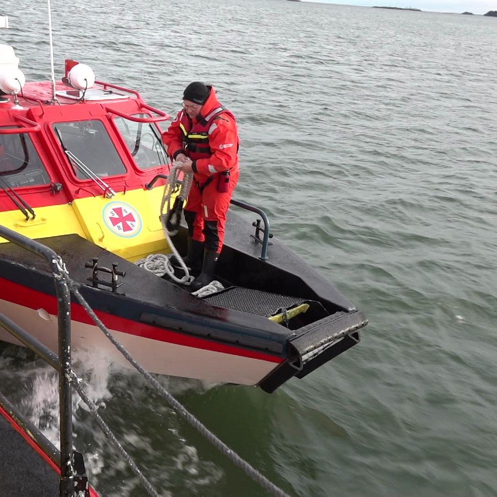 En räddningsbåt med röd och gul färg närmar sig en annan båt på vattnet. En medlem av räddningsteamet iförd röd utrustning förbereder sig för att gå ombord. Omgivningen visar lugna vatten och en klar himmel.