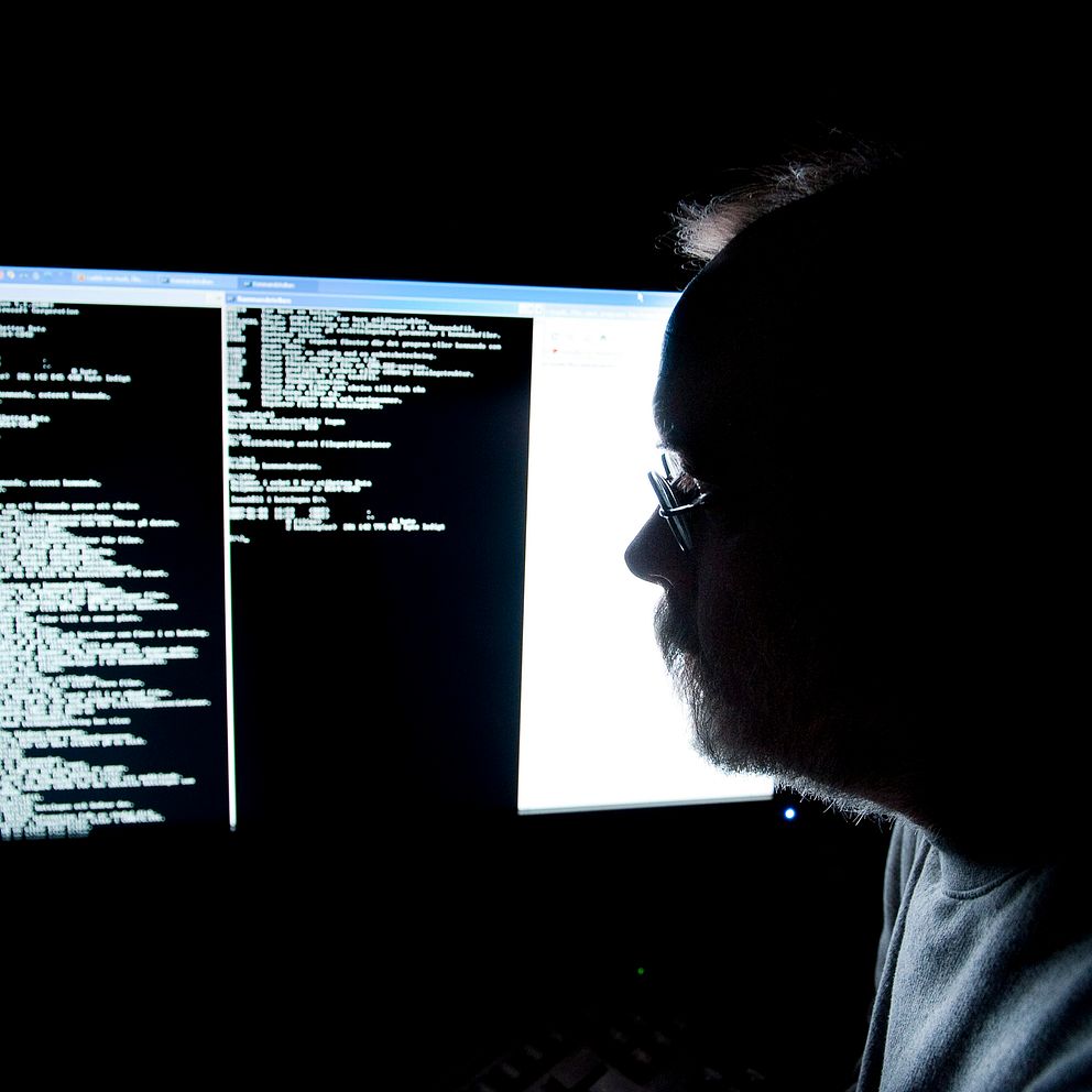 En person som sitter i mörker framför två datorskärmar, där vänster skärm visar kod i svartvit text och höger skärm har ett ospecificerat dokument eller färgglada fönster.