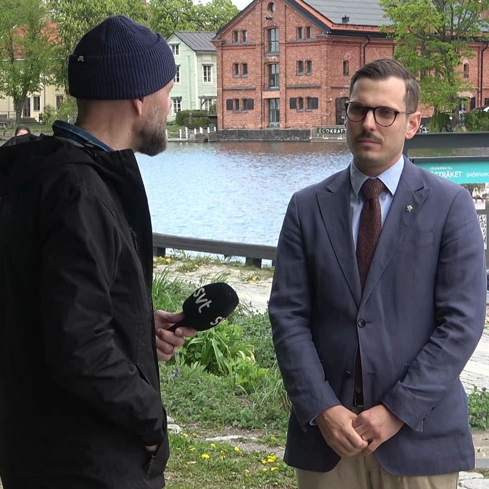 Reporter intervjuar man i kostym vid vattnet med byggnader i bakgrunden.