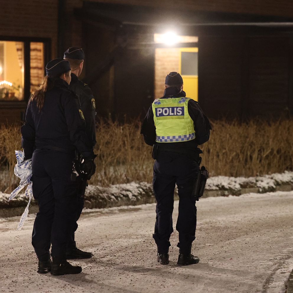 Tre poliser står på en snötäckt väg utanför en bostad, med blicken riktad mot ett hus som har ett ljus tändt i fönstret. Det är kväll och omgivningen är svagt belyst. Poliserna är klädda i uniformer och har synliga polisbrickor.