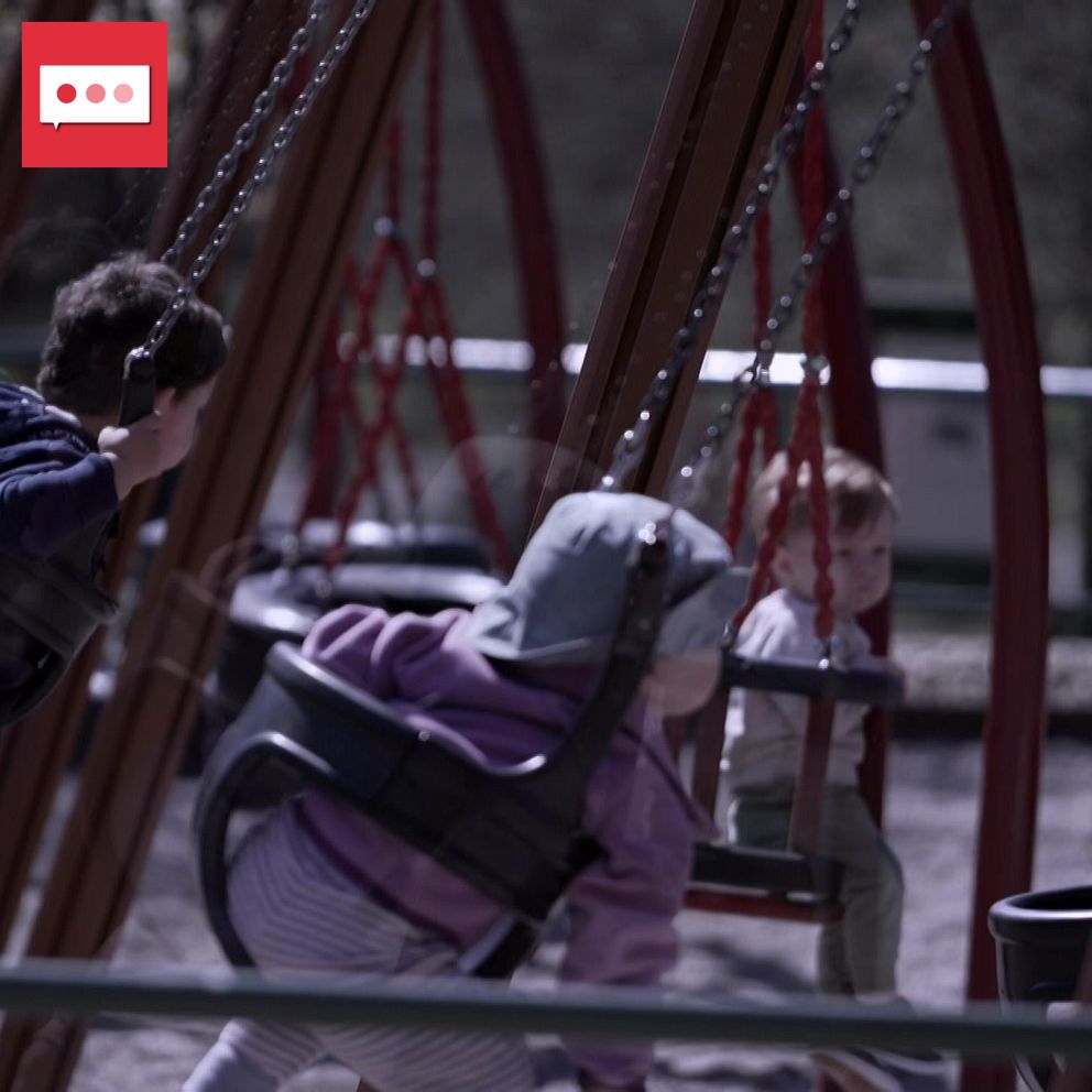 Tre barn leker på en gungställning i en park. Två av dem gungar, en pojke i en svart tröja och en flicka i gråa kläder, medan ett tredje barn sitter stilla på en gunga, klädd i ljusgrå kläder. Bakgrunden visar en grönskande miljö med träd.