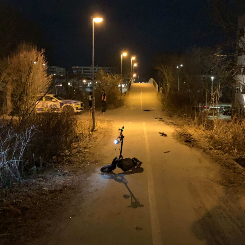 En öde cykel- och gångväg belyst av gatulampor i mörkret. På vägen ligger en elsparkcykel, medan en polisbil syns i bakgrunden. Omgivningen är omgiven av buskar och grönska, med fler byggnader i distans.