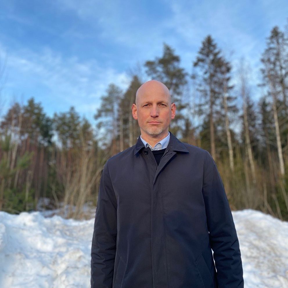 En person står framför en snötäckt mark med träd i bakgrunden under en klar himmel. Personen är klädd i en mörk jacka och skjorta, vilket ger en känsla av vinter och utomhusaktiviteter.