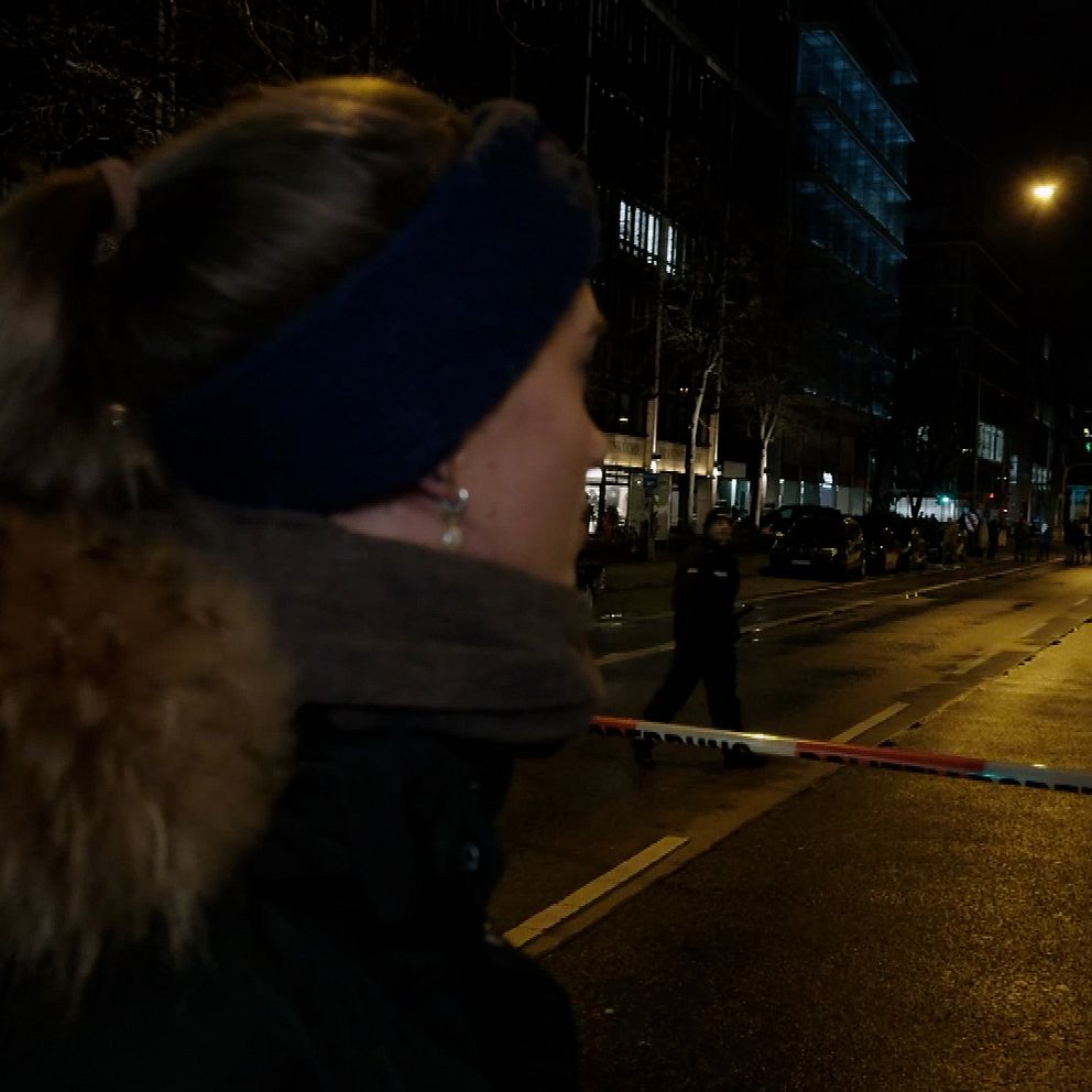 En kvinna i vinterkläder med pälsdetaljer står på en avspärrad gata på natten. Bakgrunden visar upplysta byggnader och gatubelysning längs den tomma vägen.