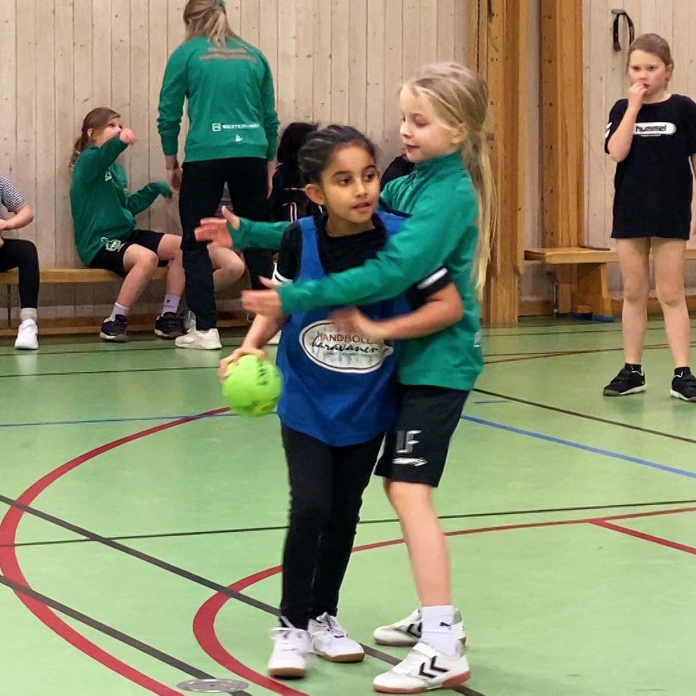 Åttaåriga Gelila spelar handboll tillsammans med sitt lag i en idrottshall. I bakgrunden sitter flera spelare på en bänk.