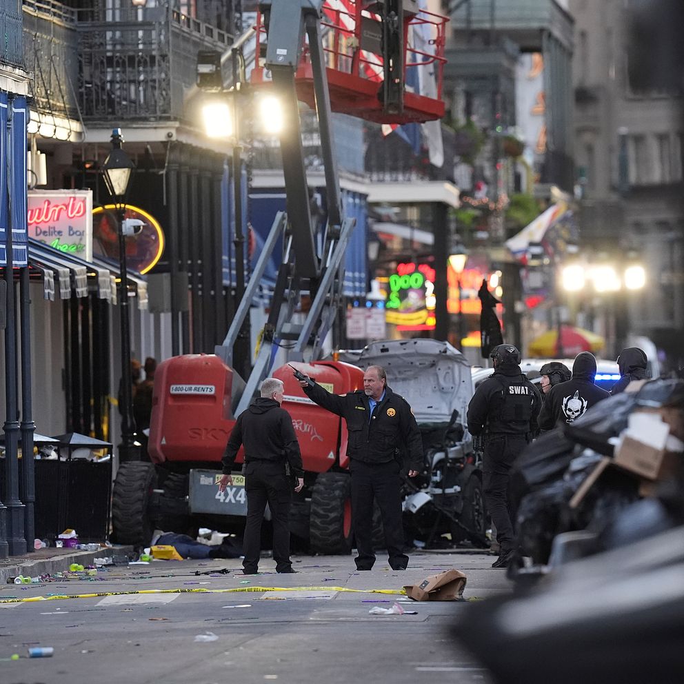 Poliser står på en gata i New Orleans, omgiven av avspärrningar och skräp efter attacken. I bakgrunden syns ett byggarbetsfordon och flera nöjeslokaler, medan den tidiga morgonen ger en dämpad belysning över scenen.