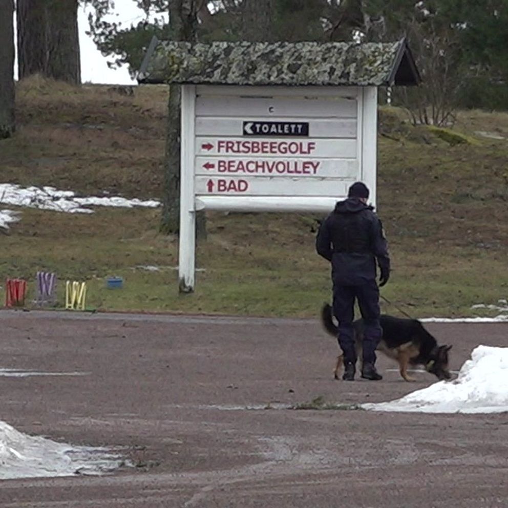 En polis går med en hund på en snötäckt väg bredvid en stor skylt som visar vägbeskrivningar till frisbeegolf, beachvolley och bad på en vintrig dag.